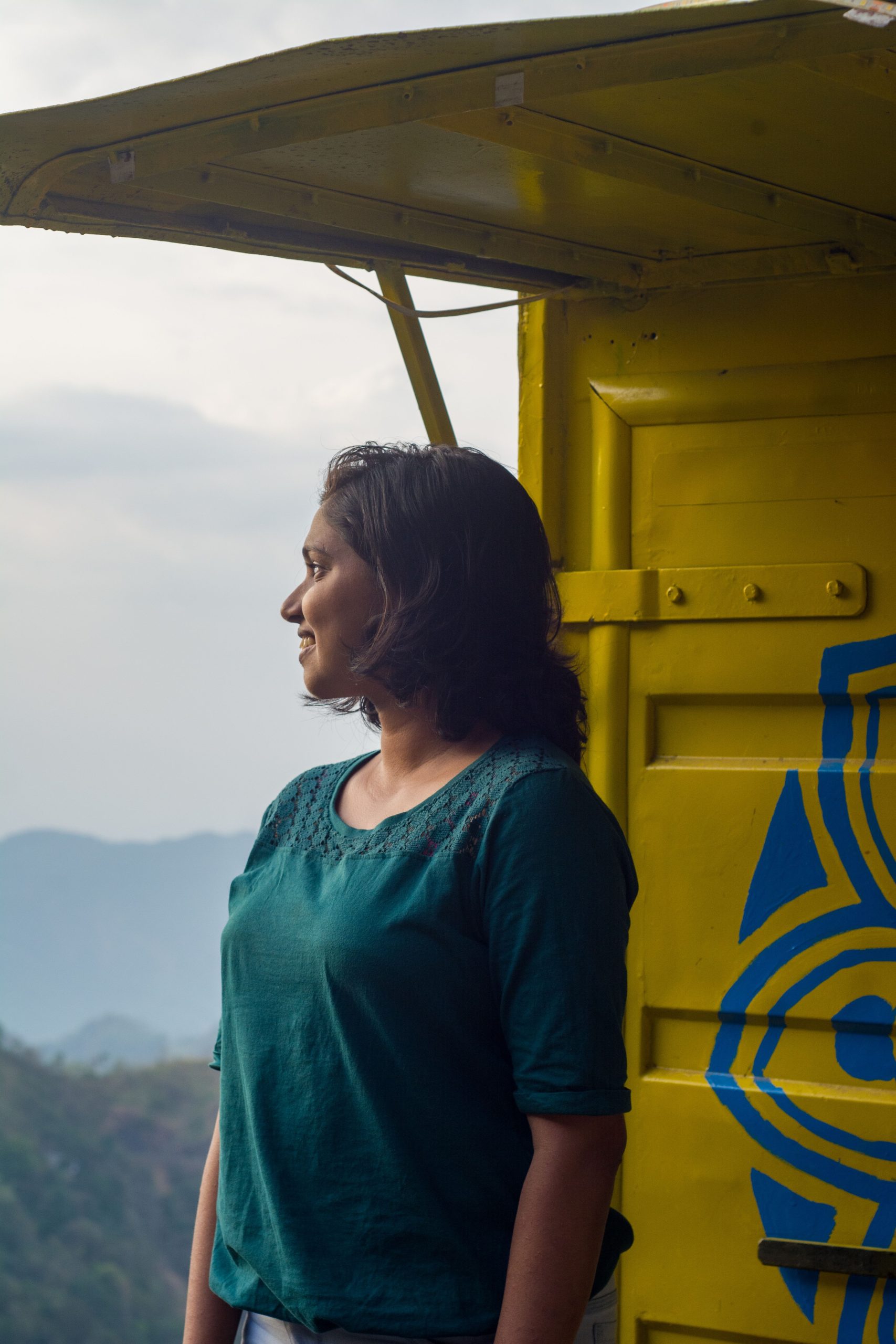 Woman in teal blouse stands by a colorful backdrop, gazing at serene mountains in India.