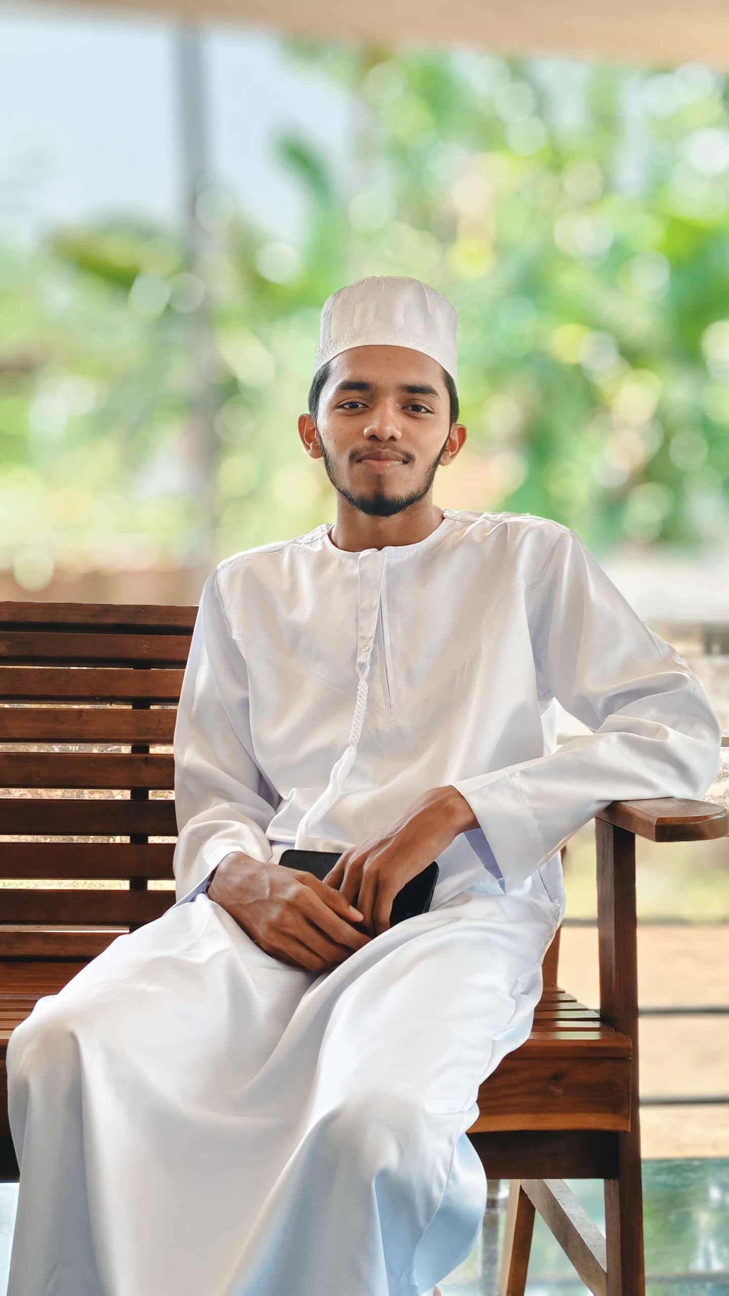 A young man in traditional attire sits on a wooden bench in a lush outdoor setting.