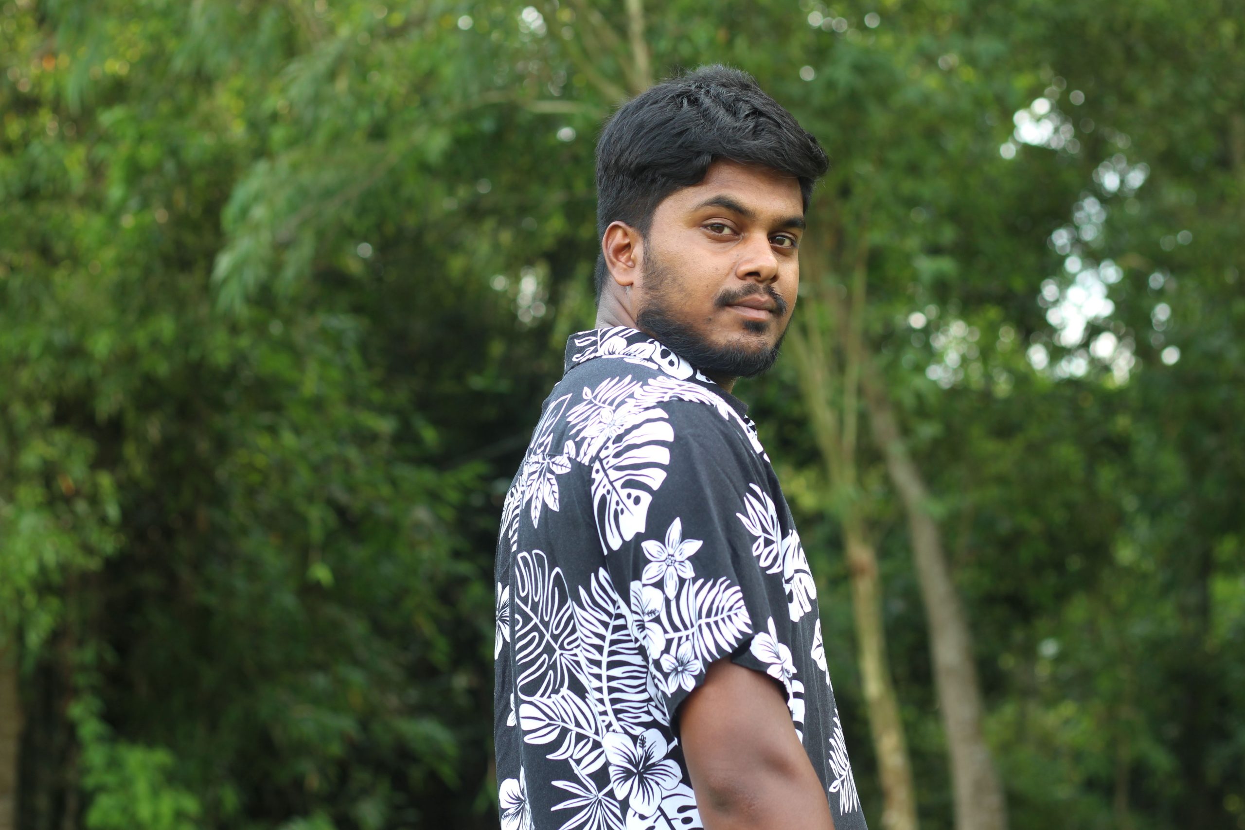 A young man wearing a floral shirt stands confidently outdoors amidst lush greenery.